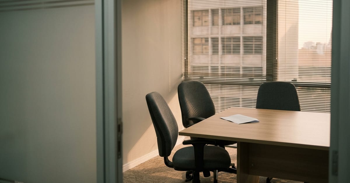 Sala de reunião de empresa com mesa vazia, cadeiras afastadas e janela com persianas